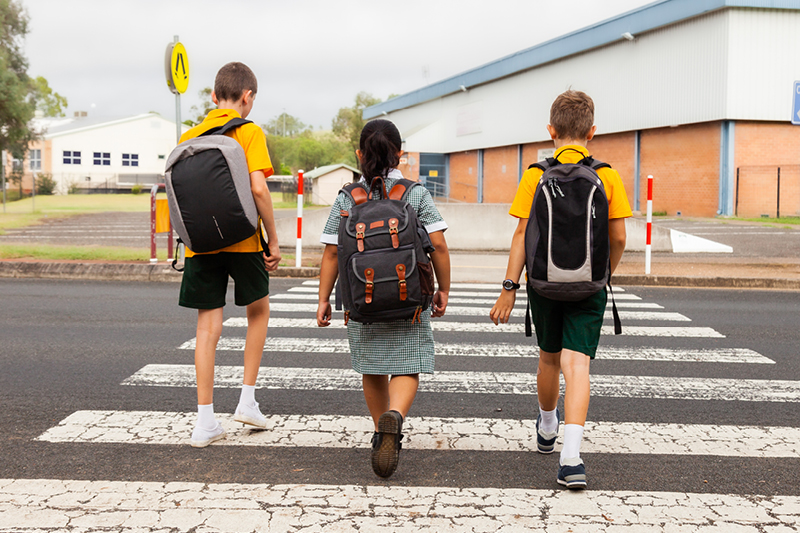 Three young children in school uniforms with backpacks on using a pedestrian crossing to get to school