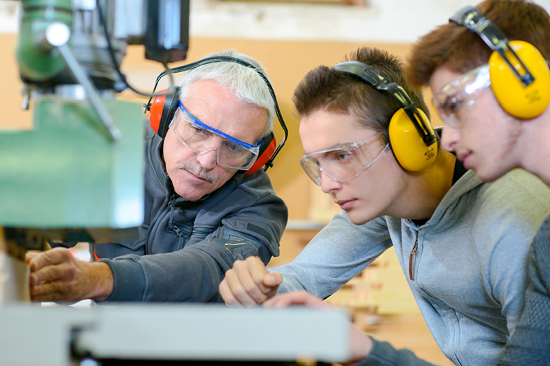 A teacher demonstraiting how to use workshop tools to two apprentices wearing safey glasses and hearing protection