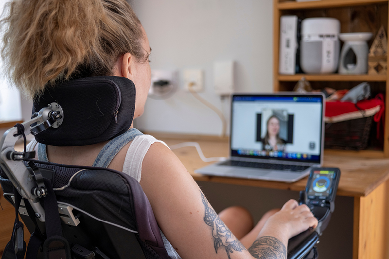 A young person in a specialised wheelchair using chair controls and laptop to engage with people online