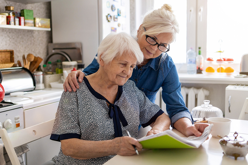 A carer assisting an elderly person with documentation, sitting in a chair in a kitchen