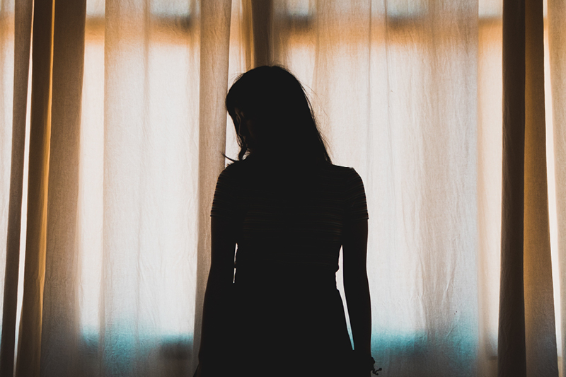 Silhouette of a person in a dark room looking down in front of closed curtains