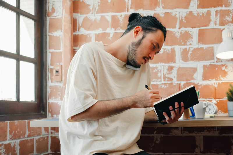 A young person with dark hair and a white shirt, sitting down at a table, holding their phone between their shoulder and cheek, writing with a pen in a notepad